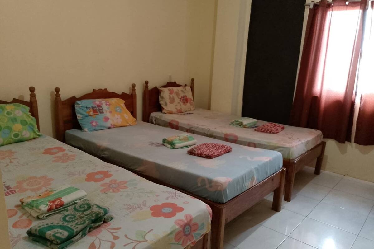 Una habitación con tres camas individuales, cada una con colcha y almohadas estampadas con motivos florales. Se ve una ventana con cortinas a la derecha.