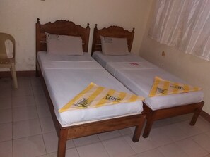 Una habitación con dos camas de madera, cada una con un colchón y una almohada blancos. También hay una silla y una ventana con cortina.