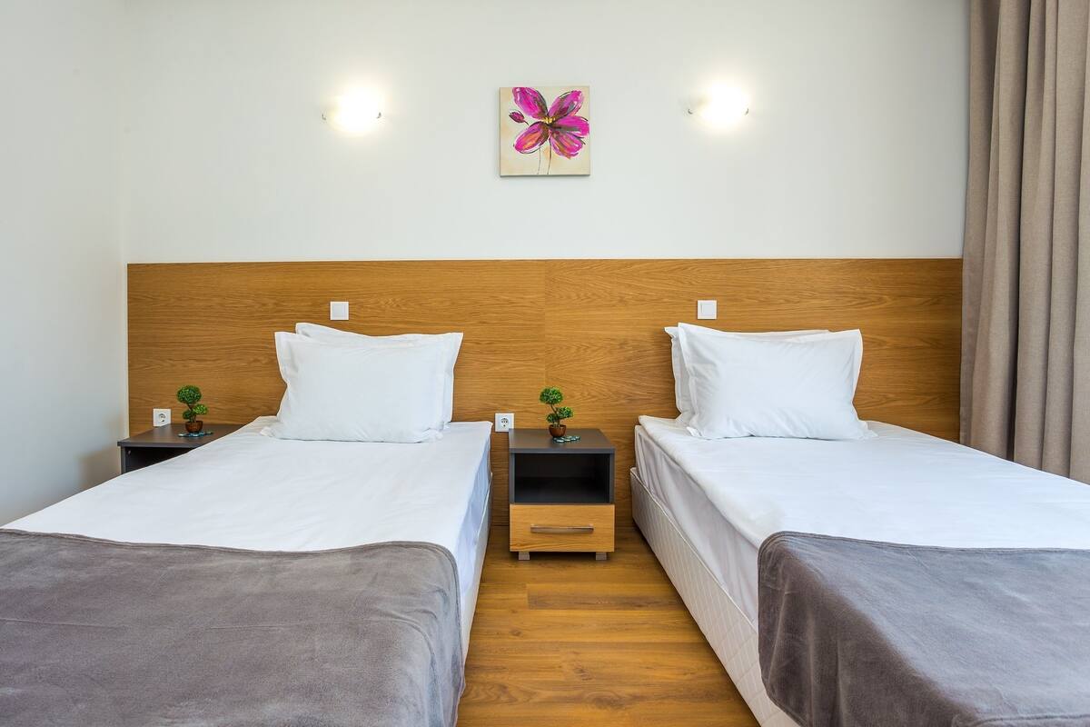 A hotel room with two single beds, wooden headboards, bedside tables with lamps, a wall-mounted light fixture, and a floral artwork.
