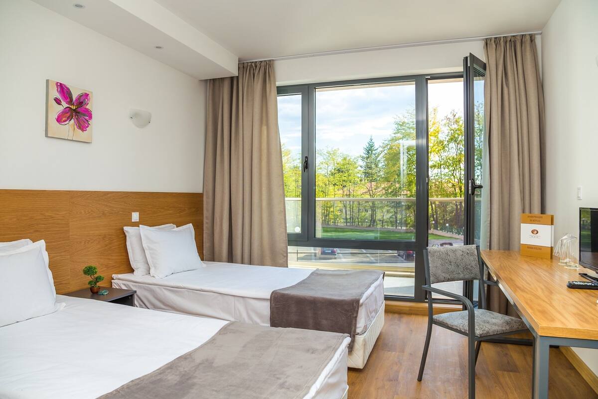 A hotel room with two beds, a wooden headboard, a desk with a chair, a window with curtains, and a view of trees.