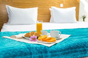 Una cama bien hecha con almohadas blancas y cabecera de madera, un edredón color turquesa, y elementos de desayuno como cruasanes, gajos de naranja y un vaso de zumo de naranja sobre una bandeja blanca.