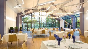 A spacious dining area with multiple tables set for guests, featuring modern lighting and large windows offering views of the outdoors. - Park Hotel Asarel (Panagyurishte)