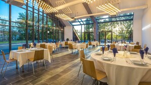 A restaurant with round tables set for dining, large windows, and hanging light fixtures. - Park Hotel Asarel (Panagyurishte)