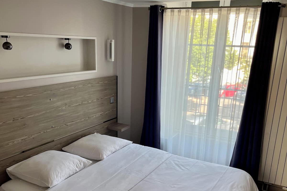Una habitación de hotel con una cama grande, un cabecero de madera, una mesita de noche y una ventana con cortinas translúcidas.