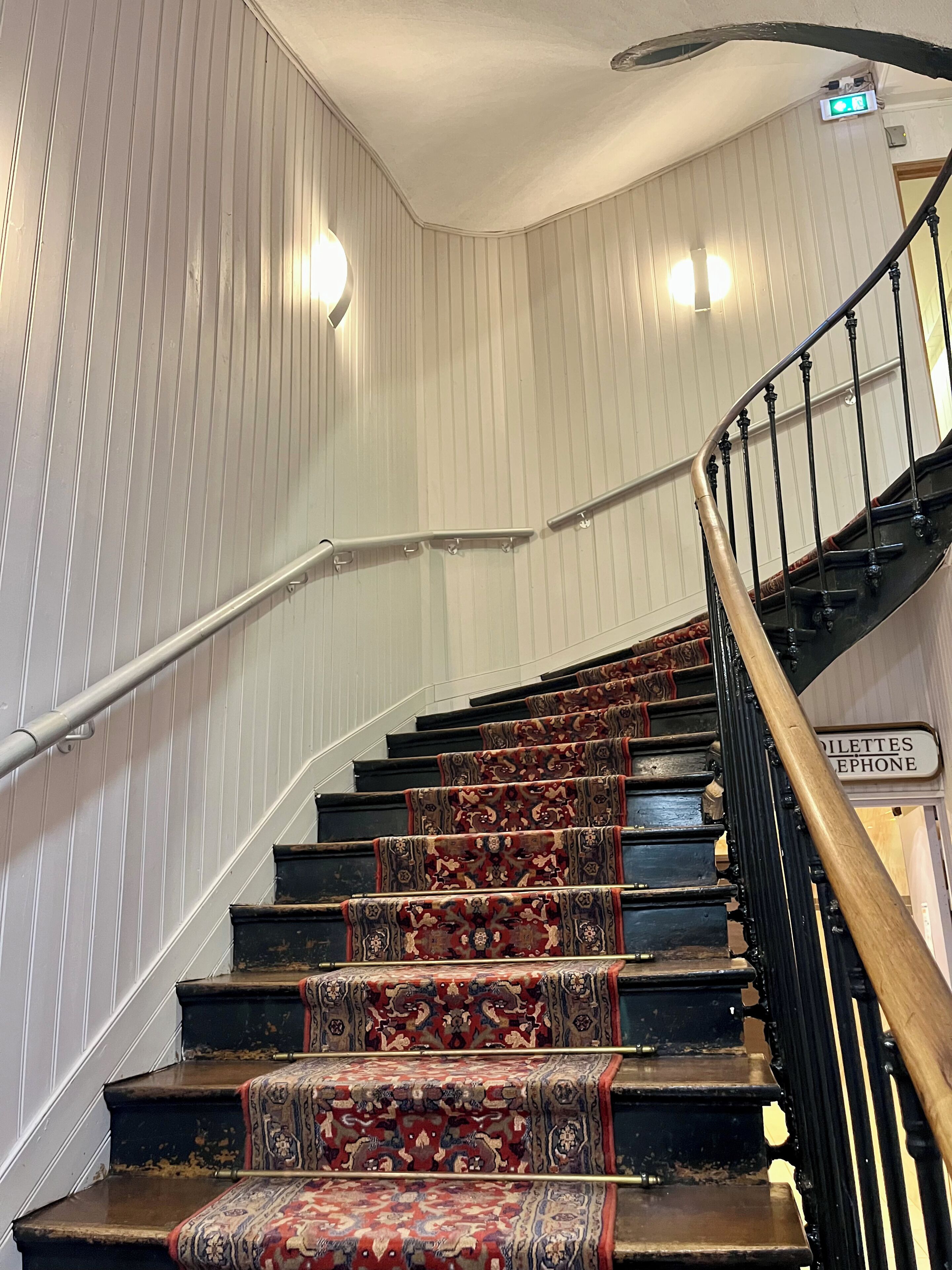 A staircase with ornate carpeted steps and a handrail, leading to a landing with a sign for the toilet and telephone.