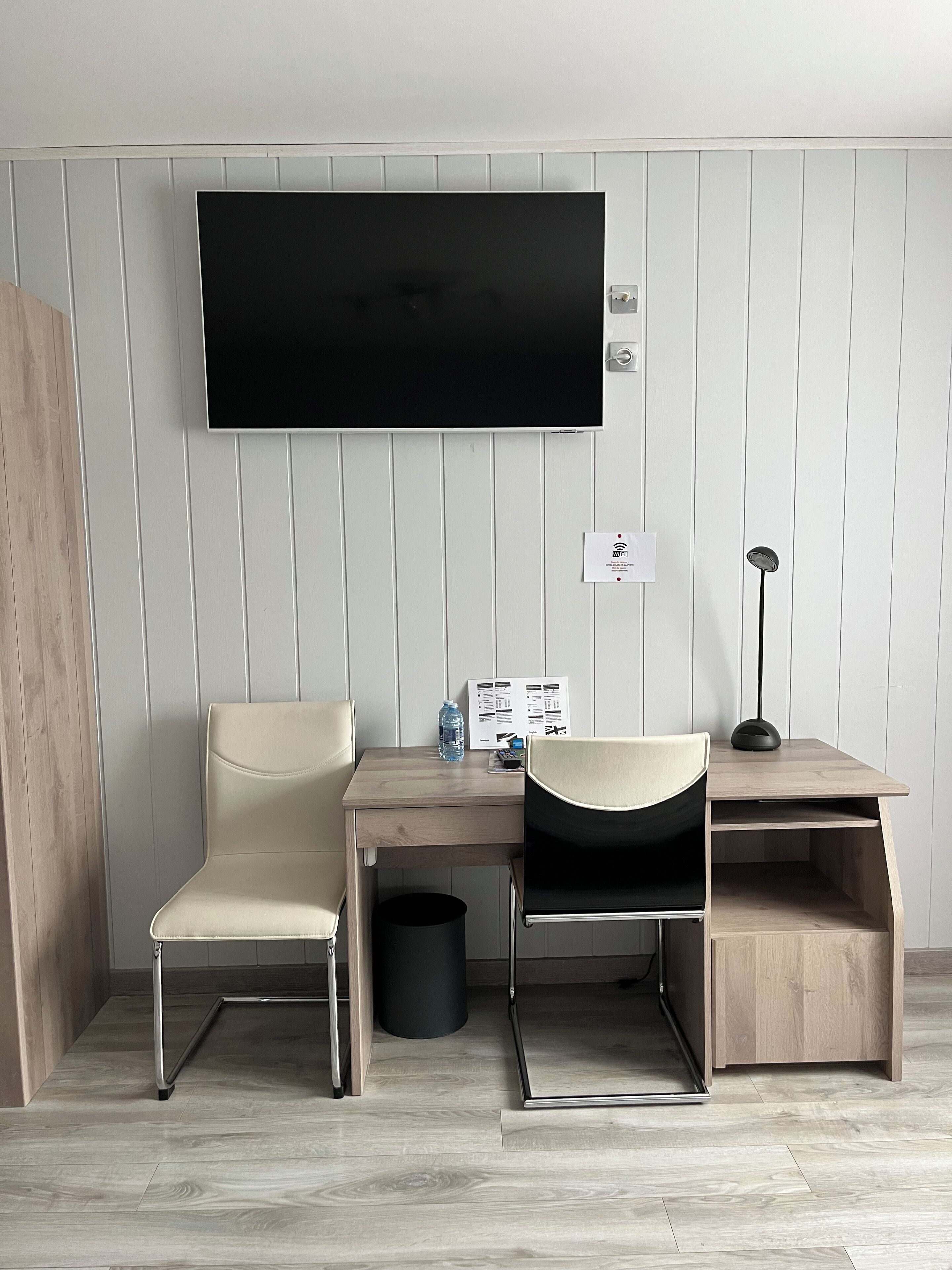 A modern hotel room with a desk, two chairs, a TV, a lamp, and a trash bin.