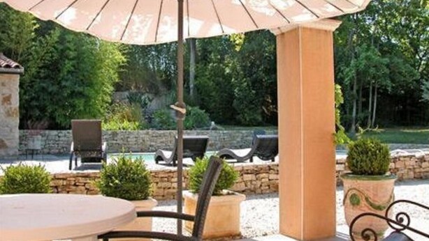 Een terras buiten met een ronde tafel, twee stoelen, een grote parasol en een zwembad op de achtergrond.