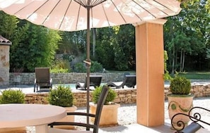Un espace extérieur aménagé en terrasse, avec une table ronde, deux chaises, un grand parasol et une piscine en arrière-plan.