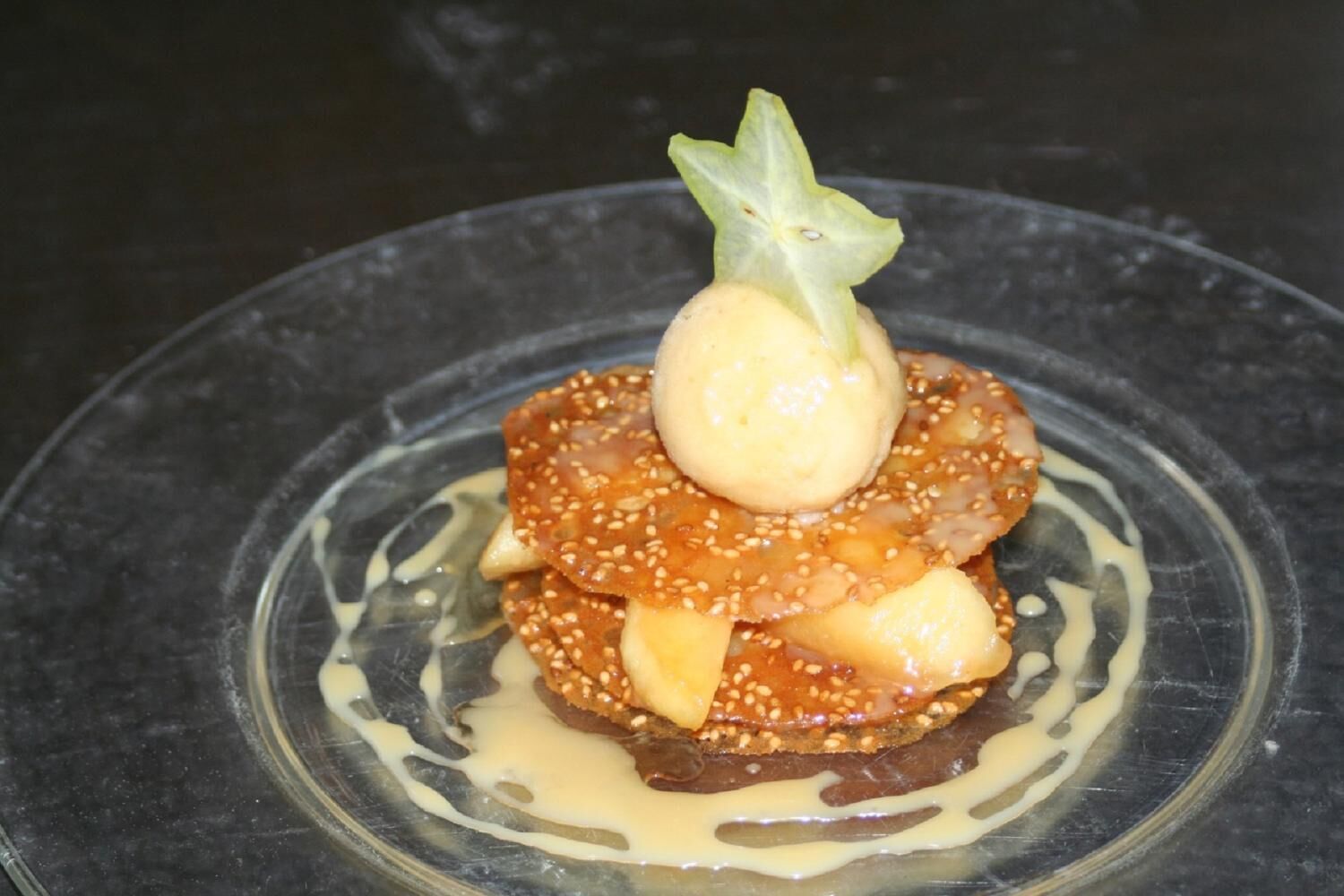 Un dessert servi sur une assiette en verre transparent, décoré d’un élément en forme d’étoile et entouré d’une sauce crémeuse.