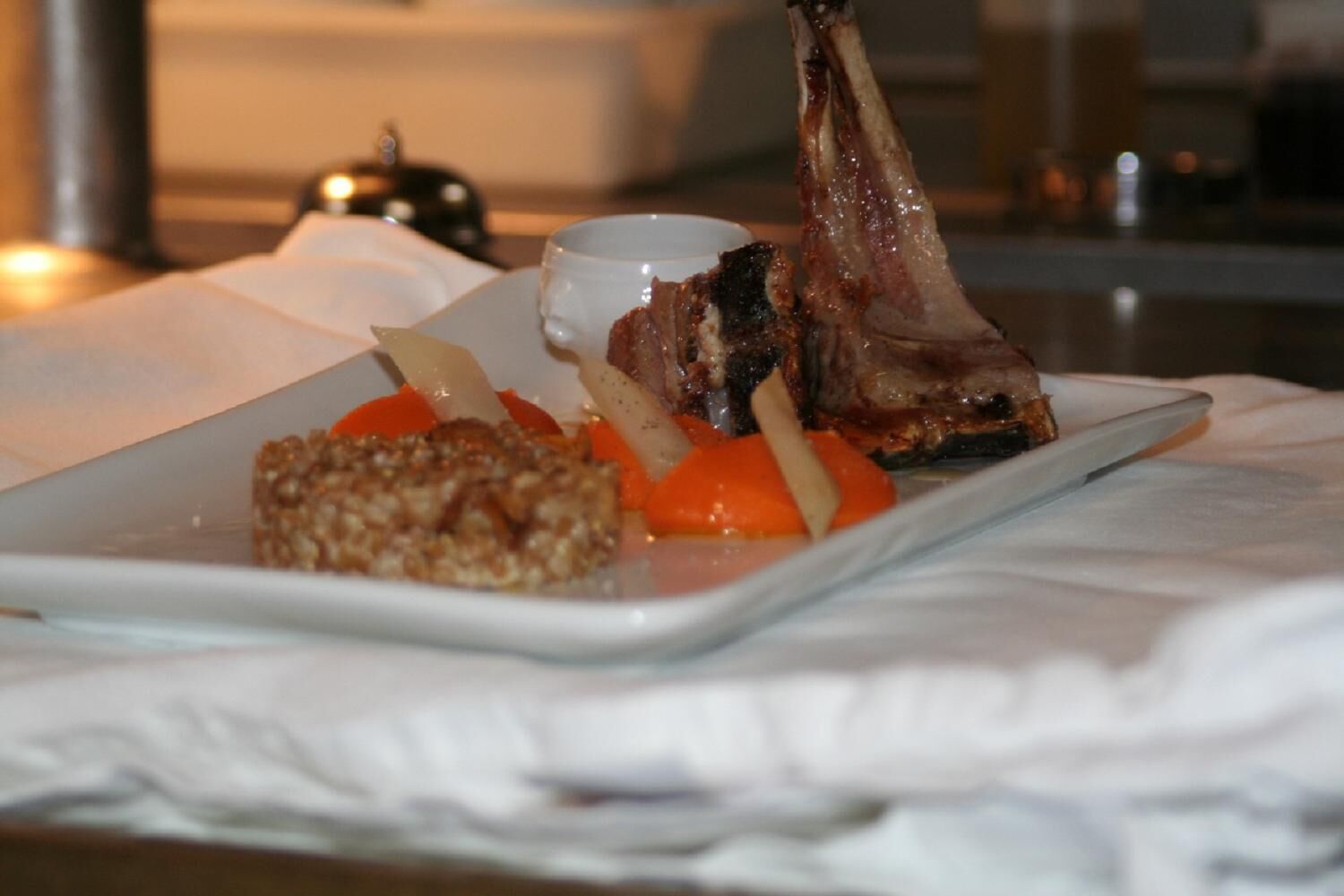 Un plato con un jarrete de cordero, zanahorias y un recipiente pequeño con salsa, posiblemente en un restaurante.