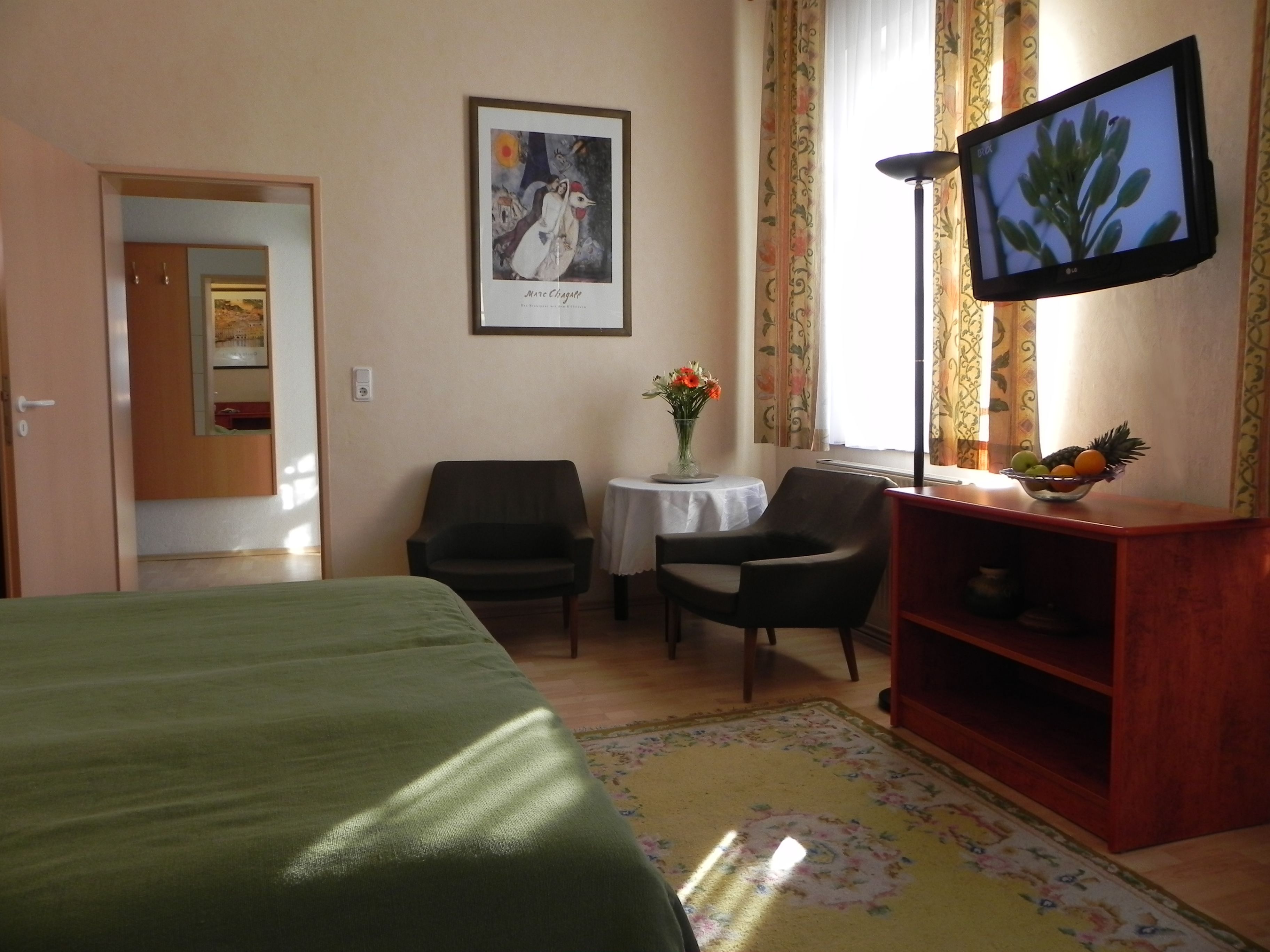 Habitación de hotel con cama, escritorio y silla, televisor, florero con flores y frutero.