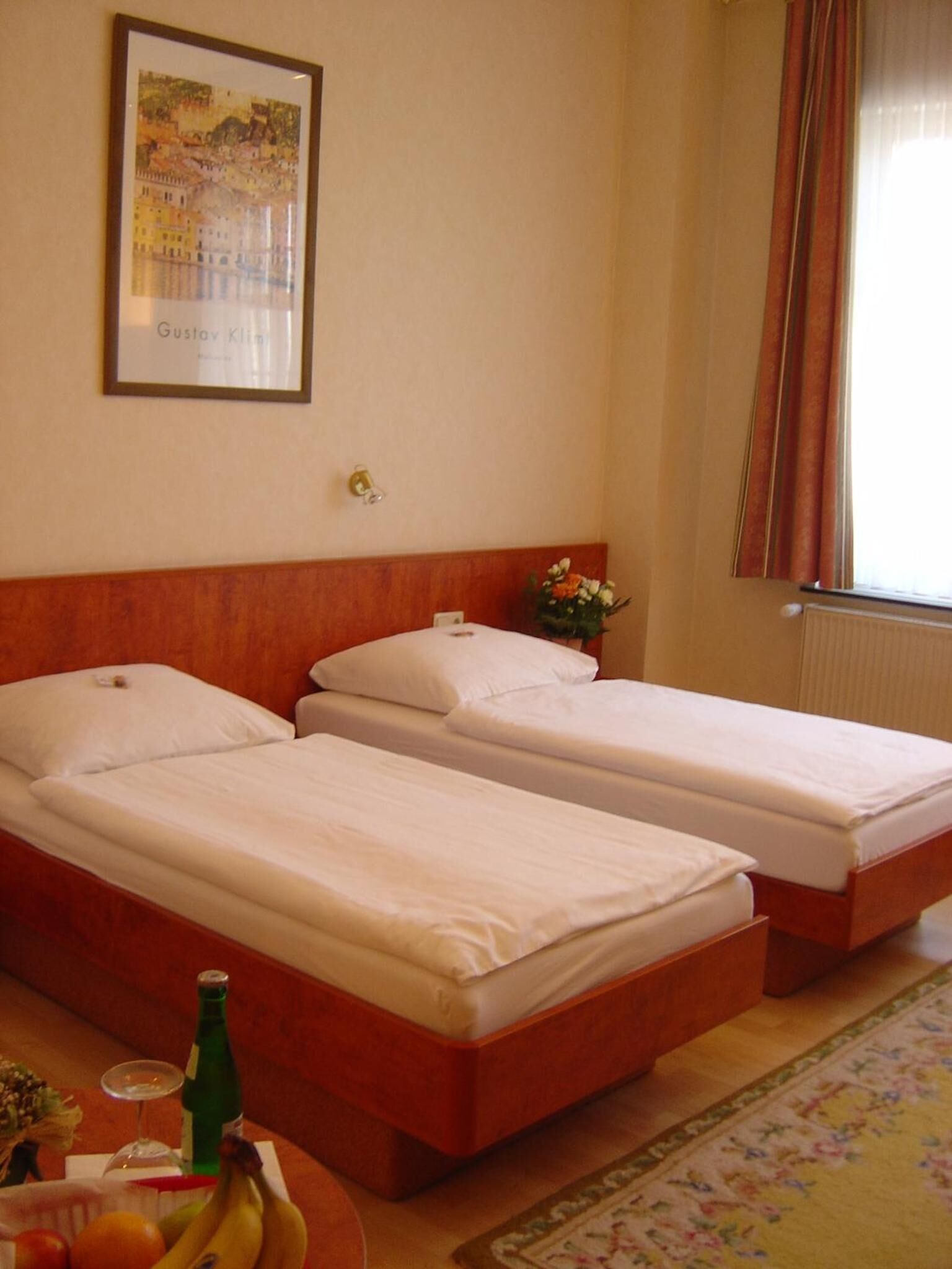 Ein Hotelzimmer mit zwei Einzelbetten, einem gerahmten Gemälde an der Wand, einem kleinen Tisch mit einer Flasche und Obst sowie einem Fenster mit Vorhängen.