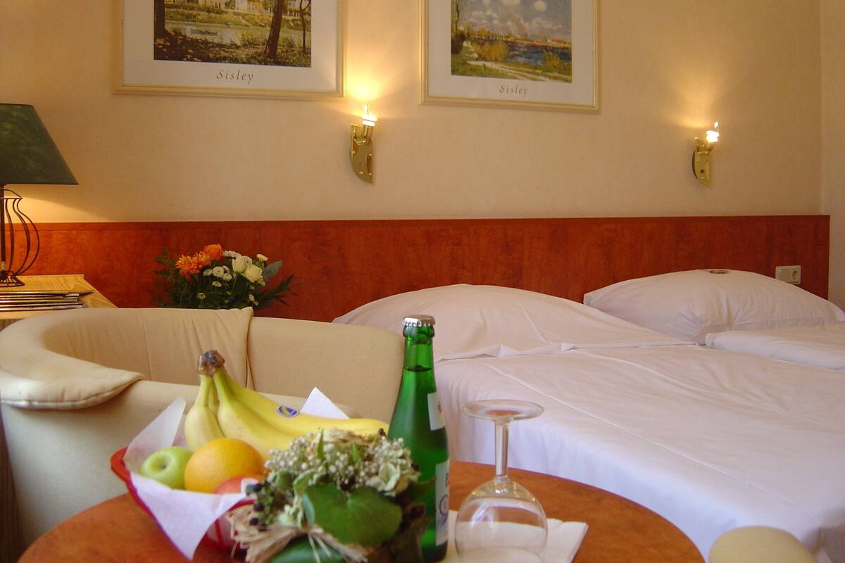 Ein Hotelzimmer mit zwei Betten, einem Tisch mit Obst und einer Flasche sowie zwei gerahmten Bildern an der Wand.