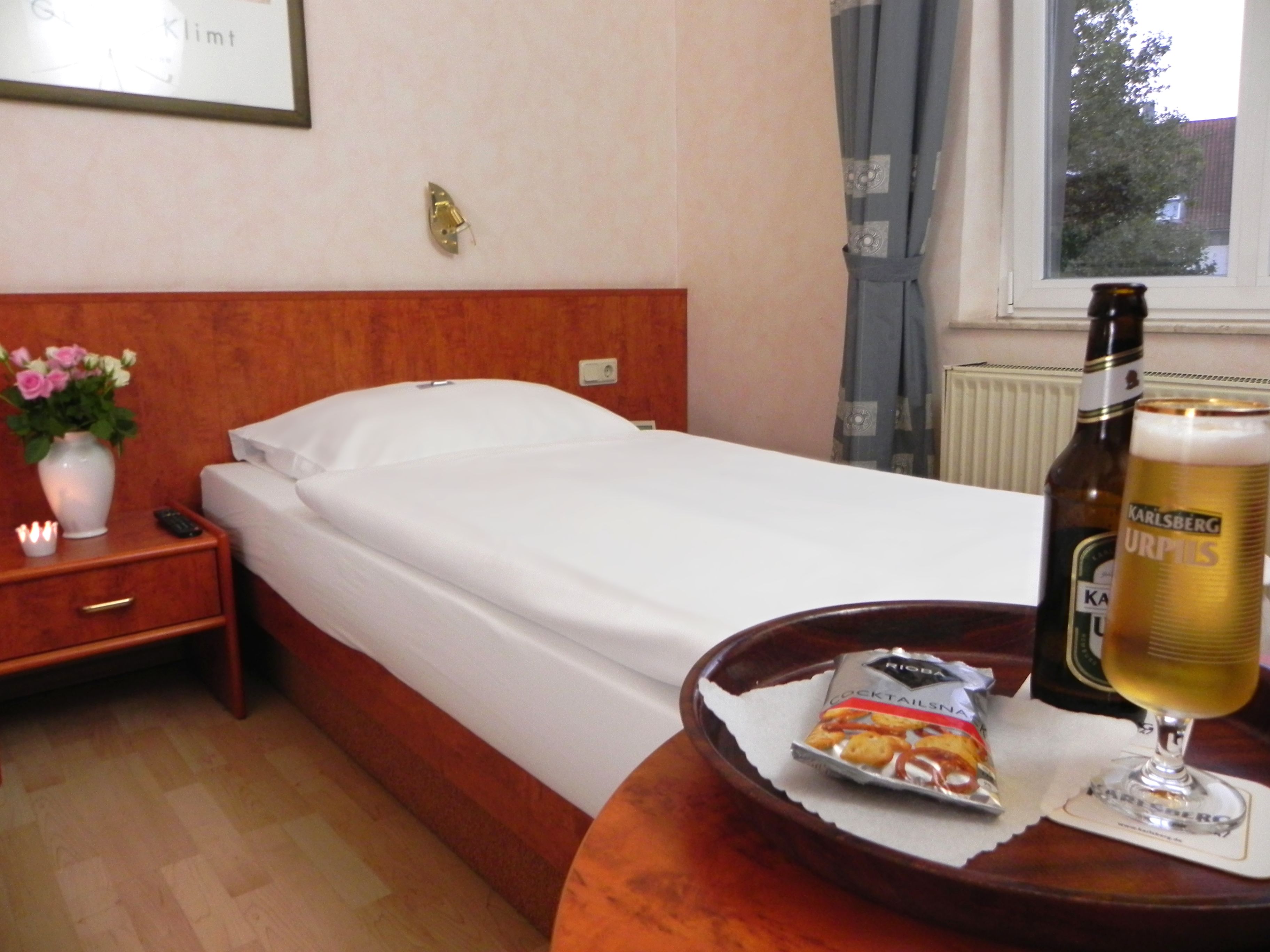 A hotel room with a bed, a nightstand with a vase of flowers, a bottle of beer, and a snack packet.