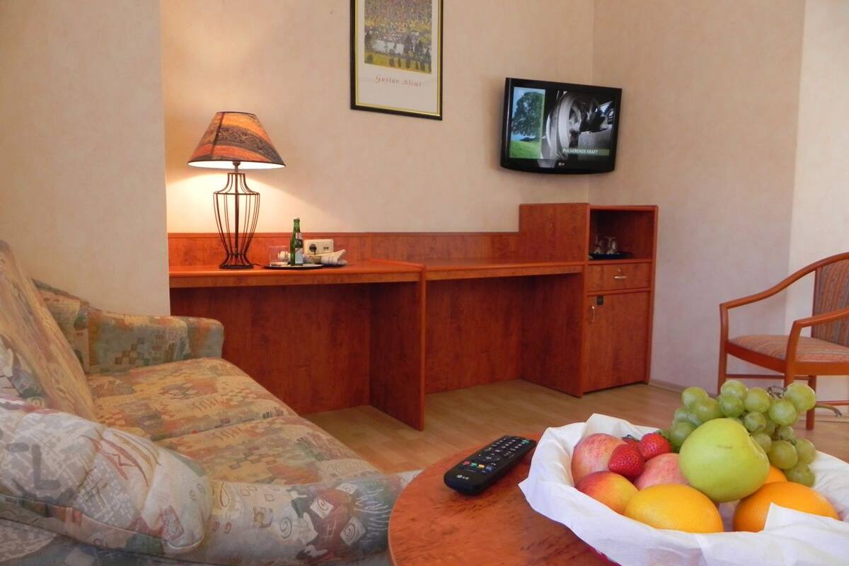 Ein Wohnzimmer mit einer Couch, einem Holztisch mit Obstschale, einer Fernbedienung, einem an der Wand befestigten Fernseher und einer Lampe.