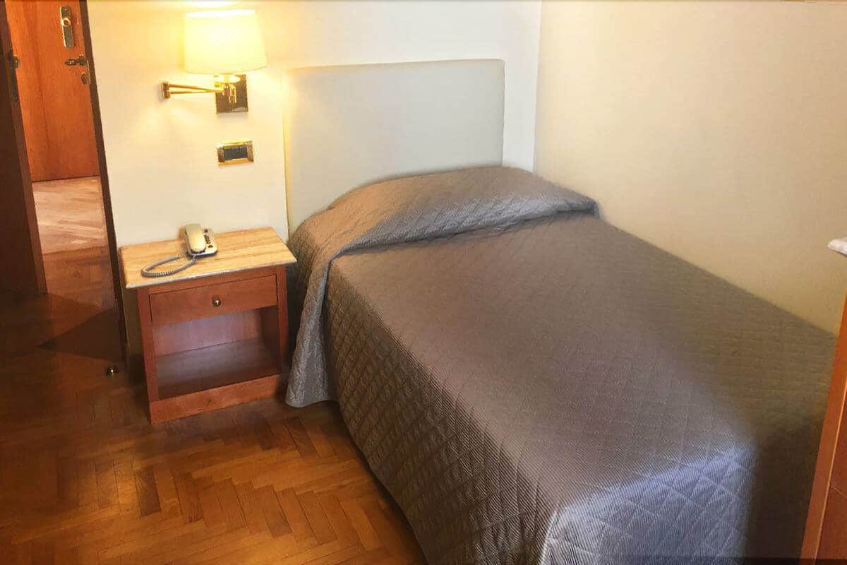 A small, single-bed room with a wooden nightstand, a lamp, and a telephone.