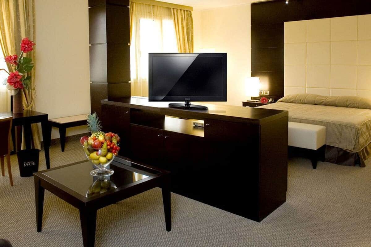 A hotel room with a bed, a TV on a desk, a fruit bowl, and a vase of flowers.