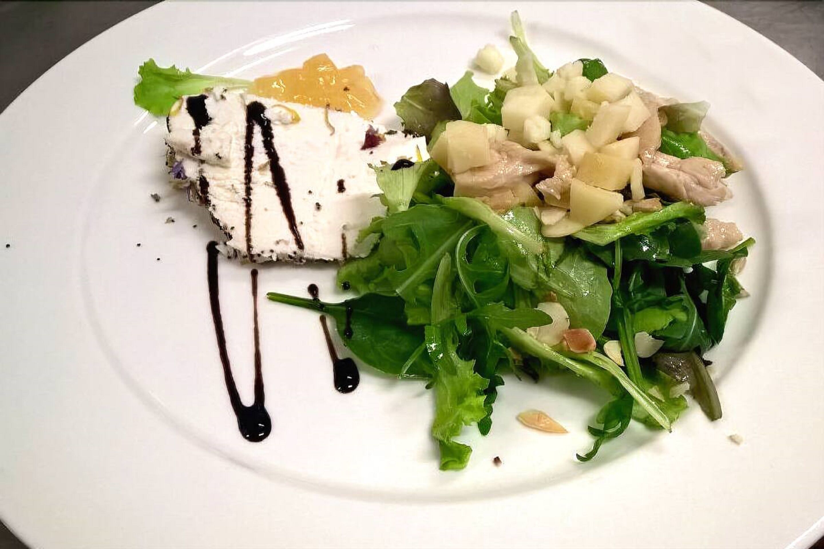 A plate with a piece of white food drizzled with black sauce, a green leafy salad, and diced potatoes.