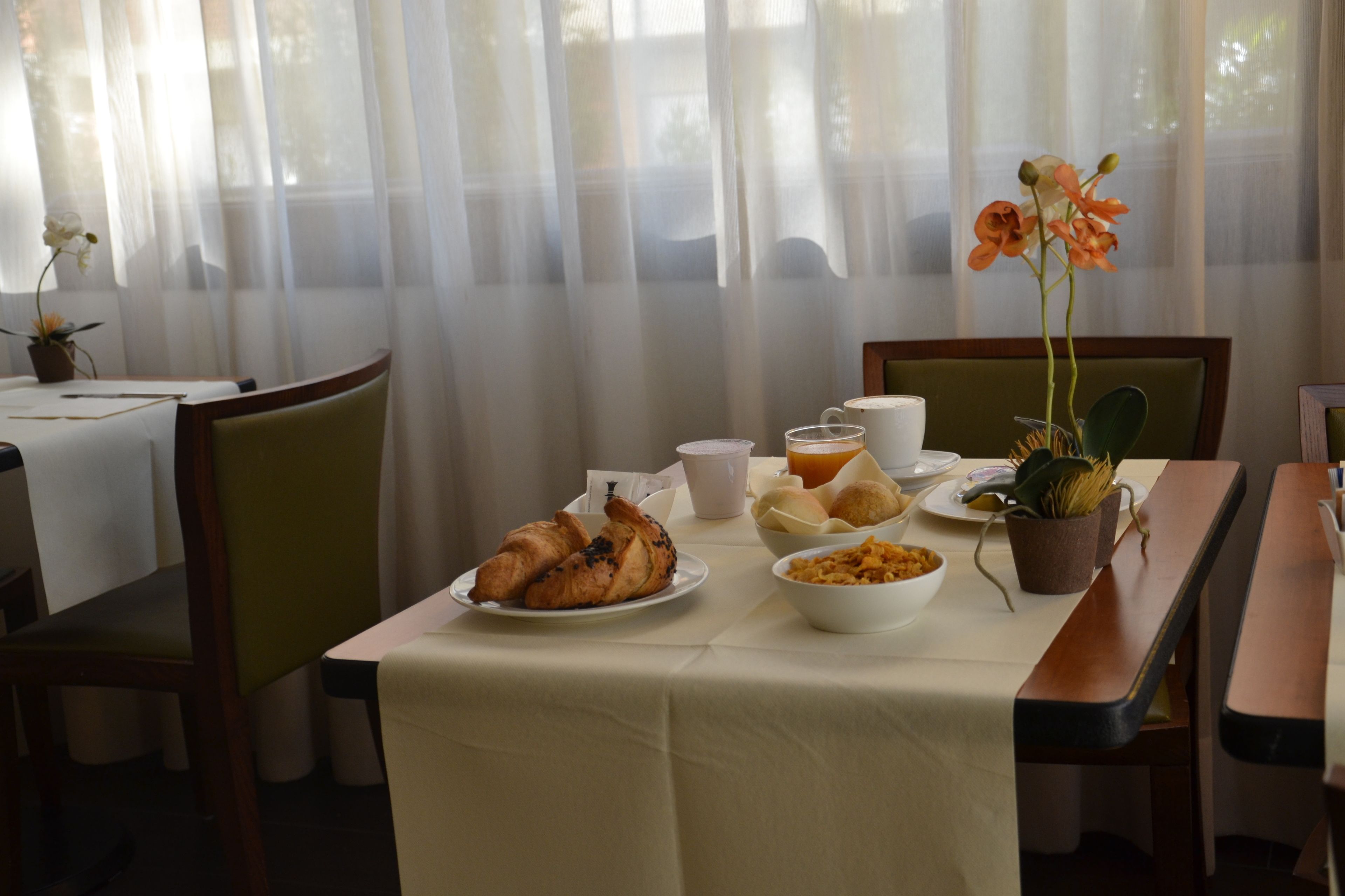 A dining area with a table set for breakfast, including croissants, bread rolls, and a bowl of cereal. There is a potted plant with flowers on the table.