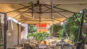 An outdoor dining area with tables and chairs under a canopy, surrounded by greenery and buildings. - Hotel Airone (Grosseto)