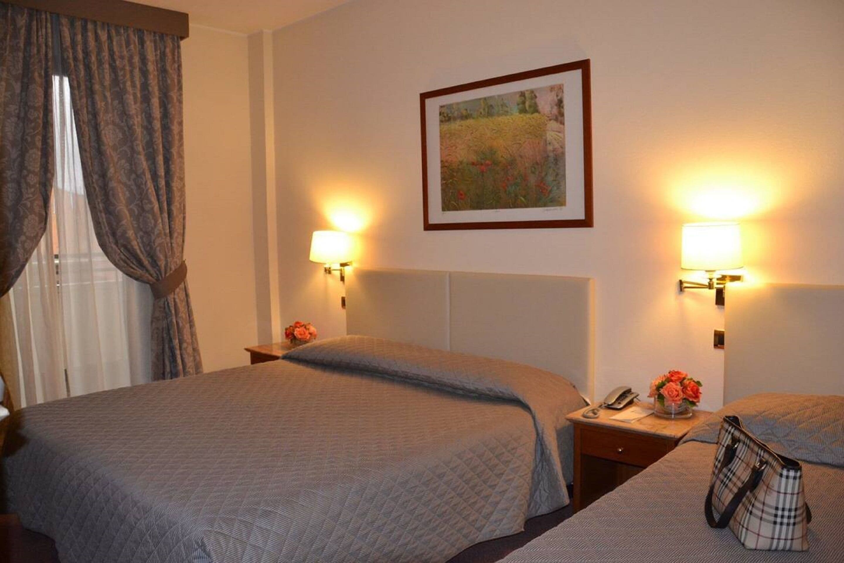 A hotel room with two beds, a nightstand with a lamp, a framed picture on the wall, and a window with curtains.