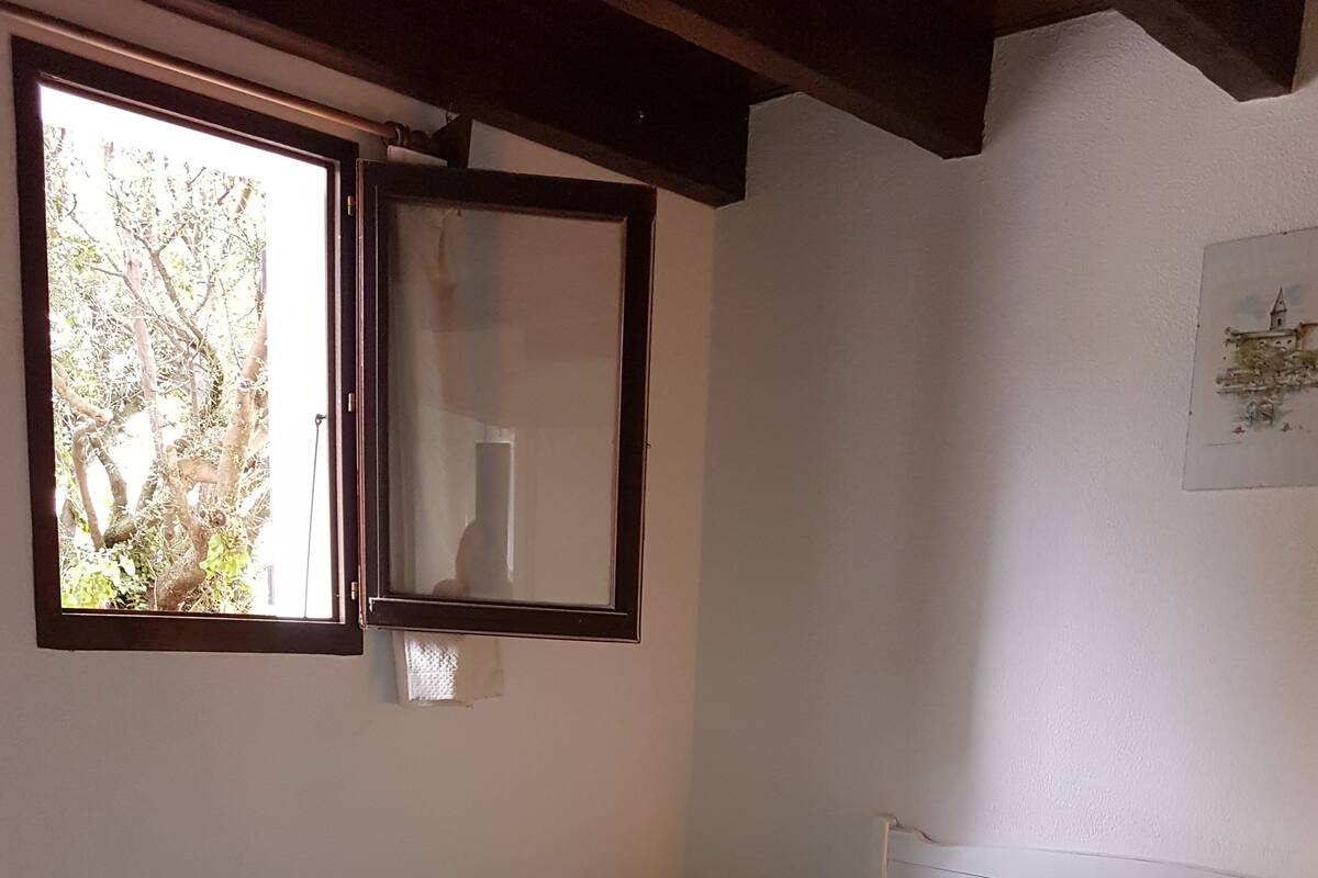A bedroom with a bed, a nightstand, a window with a view of trees, and a wall-mounted picture.