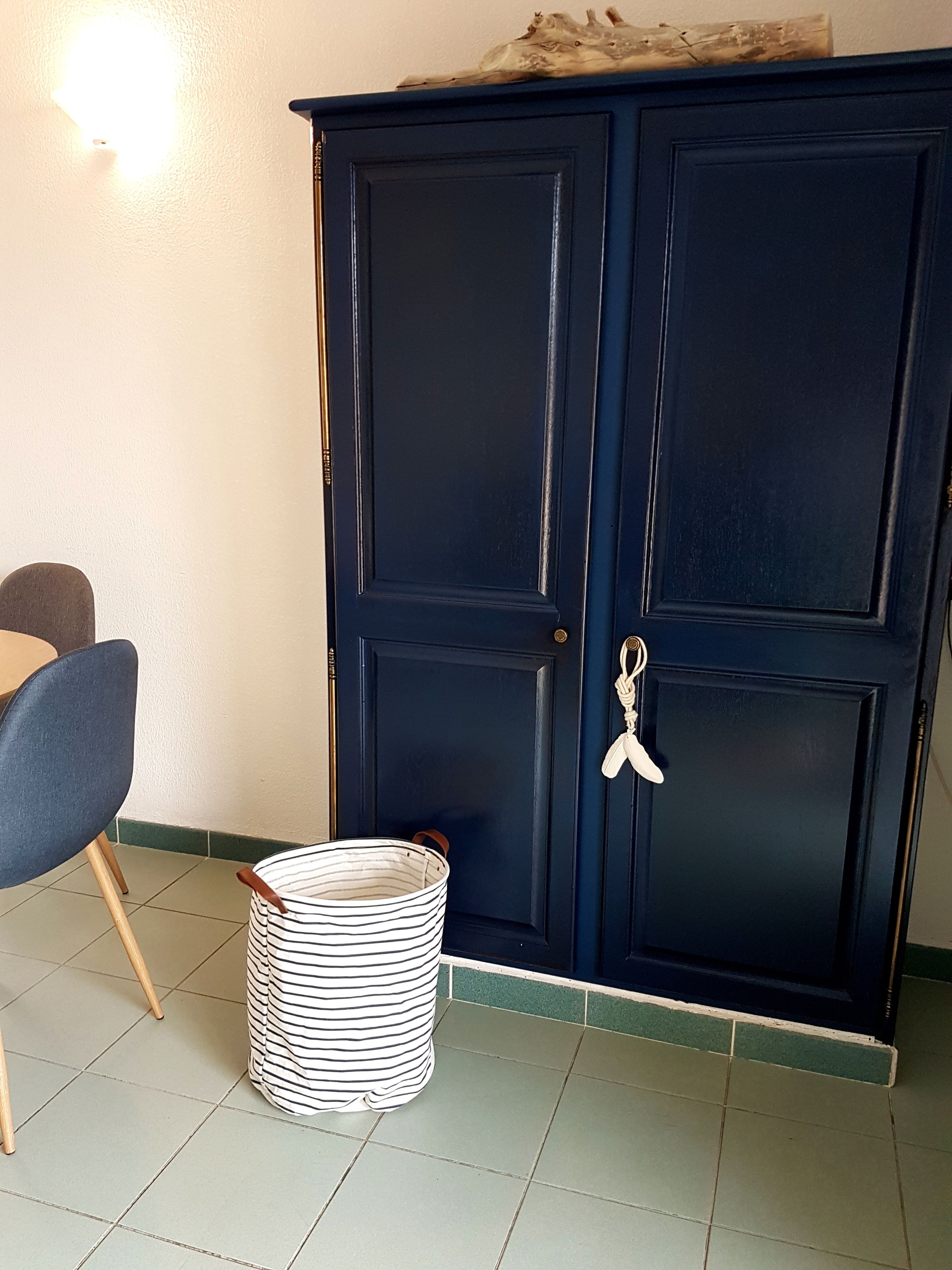 A room with a dark blue wardrobe, a striped basket, a chair, and a wall-mounted light.