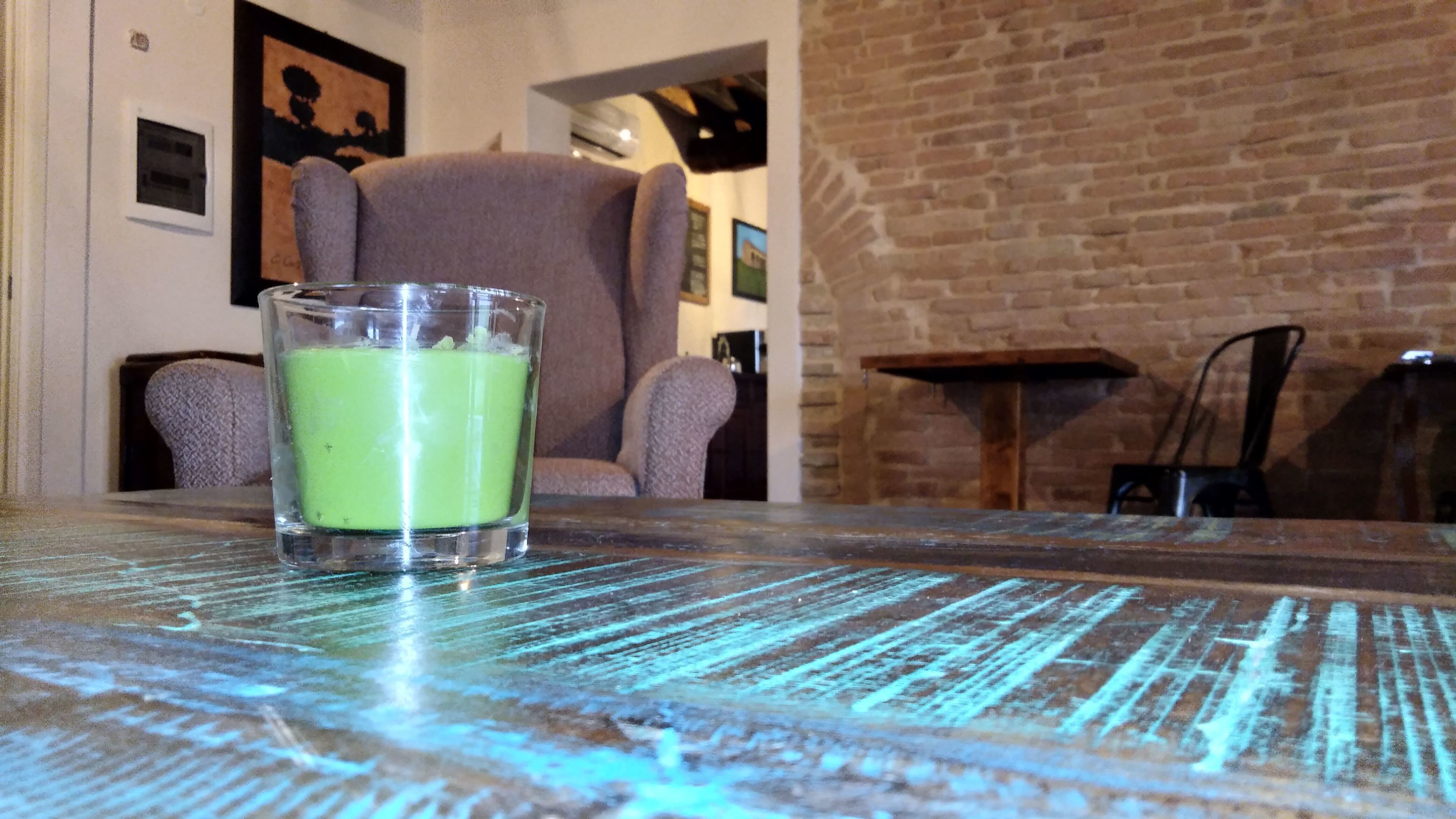 Un vaso de bebida verde sobre una mesa de madera, con un muro de ladrillo y una silla al fondo.