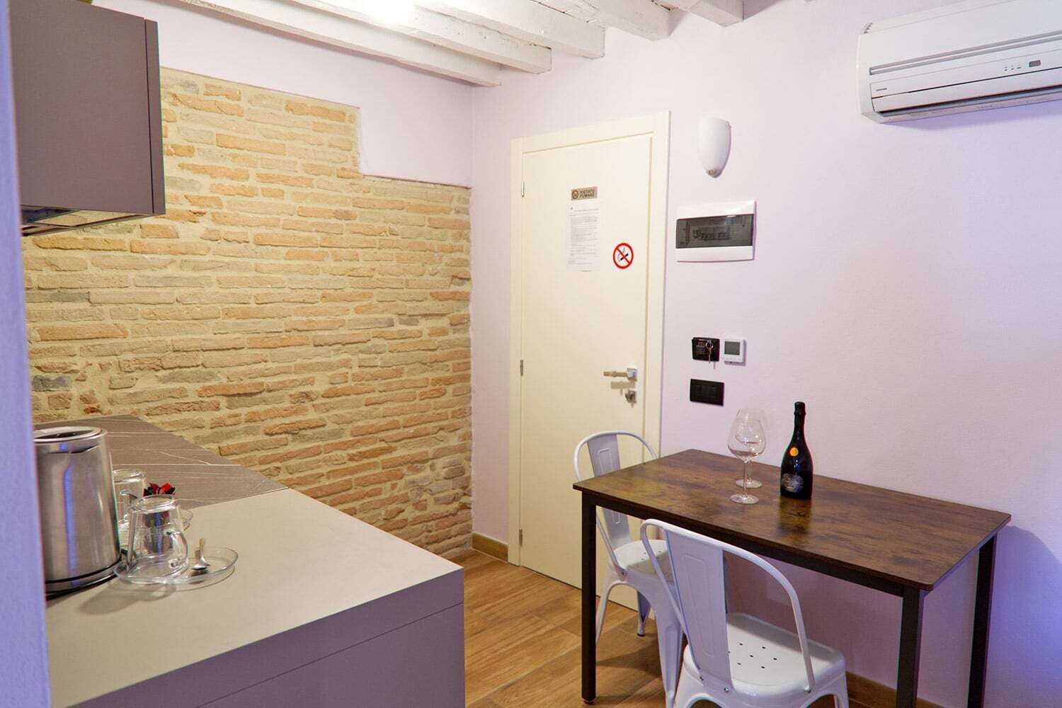 Una cucina compatta con una parete in mattoni, una porta bianca con il cartello ‘vietato fumare’, un piccolo tavolo con una bottiglia e un bicchiere, e una sedia bianca.