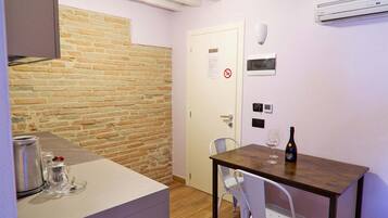 Una cucina compatta con una parete in mattoni, una porta bianca con il cartello ‘vietato fumare’, un piccolo tavolo con una bottiglia e un bicchiere, e una sedia bianca.