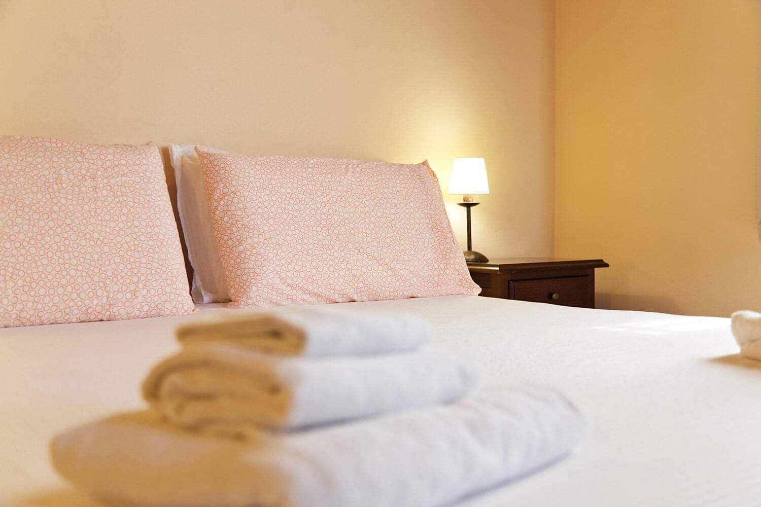 A hotel room with a neatly made bed, two pillows, a bedside lamp, and a rolled towel on the bed.