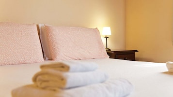 A hotel room with a neatly made bed, two pillows, a bedside lamp, and a rolled towel on the bed.