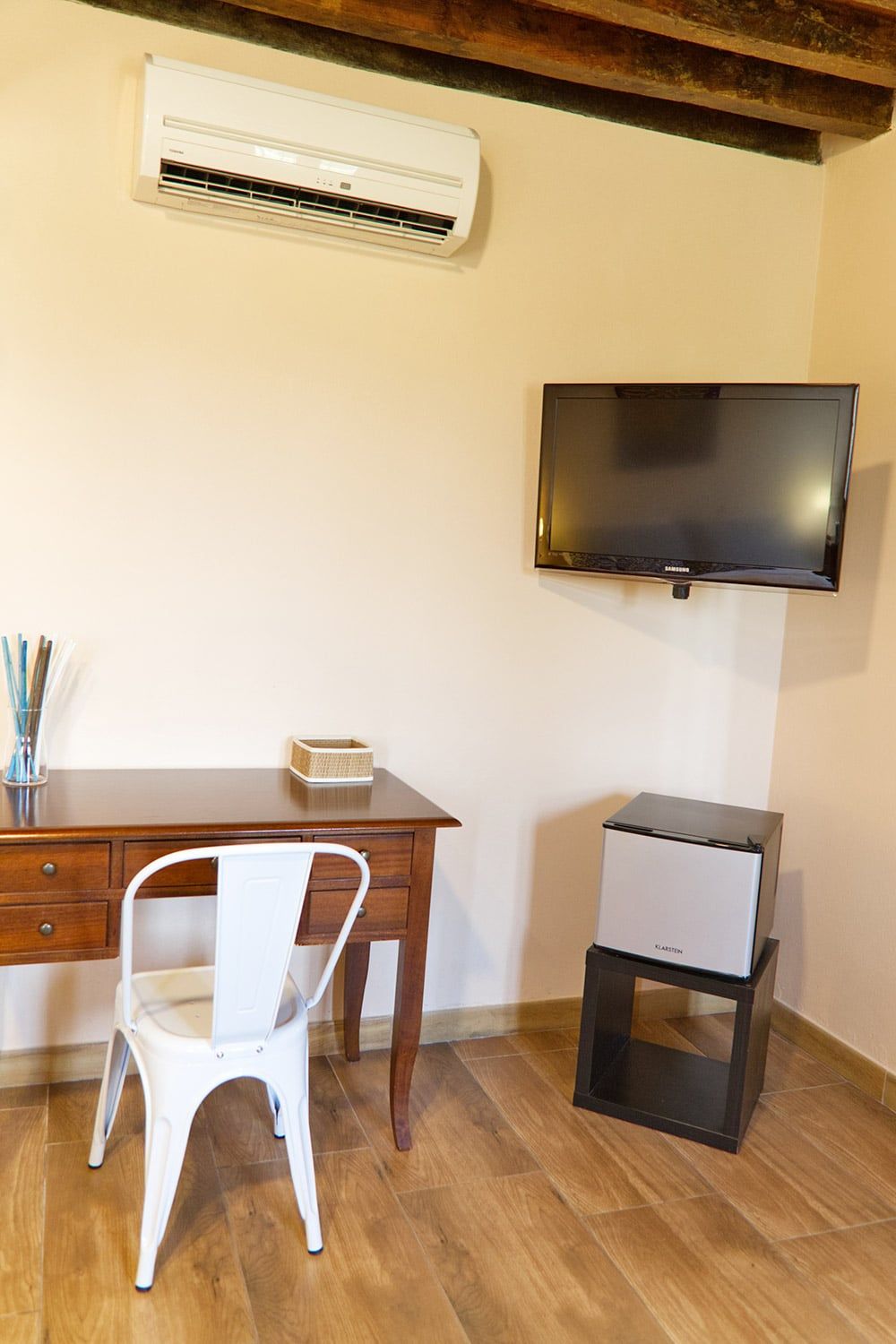 Una habitación con un escritorio de madera, una silla blanca, un televisor de pantalla plana montado en la pared y un mueble bajo.