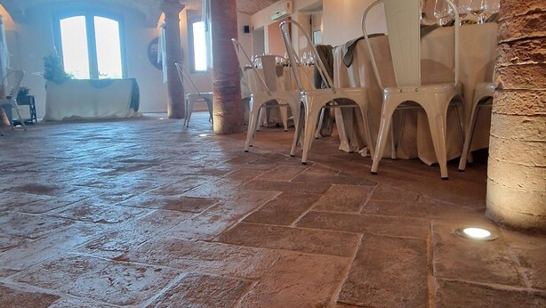 Een ruime hal met stenen vloer en bakstenen muren, met rijen witte stoelen en tafels, waarschijnlijk bedoeld voor diner of evenementen.