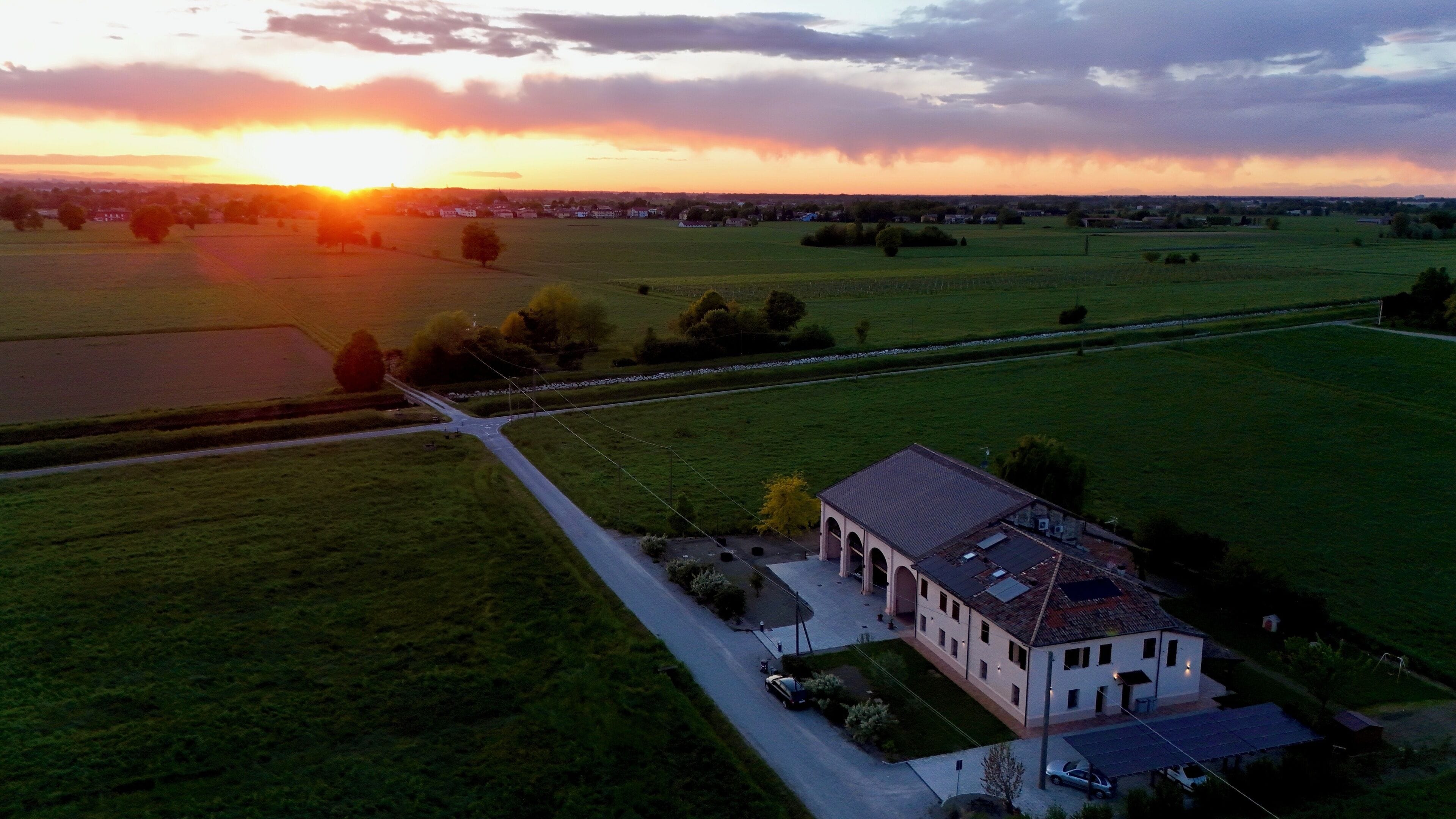 An aerial view of a rural landscape with a large house featuring a central courtyard, surrounded by open fields and a small body of water.