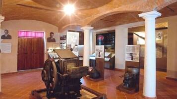 Une exposition de musée présentant des objets historiques, dont une presse à imprimer ancienne, exposés sur un sol carrelé, avec des panneaux d’information et une porte en bois en arrière-plan.