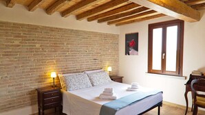 Een slaapkamer met een bakstenen muur, houten balken in het plafond, een bed met kussens, een nachtkastje, een bureau en een raam.