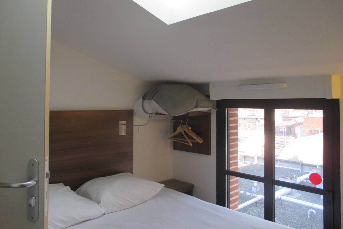 A hotel room with a bed, a wooden headboard, a window with a view of buildings, and a hanging coat rack.