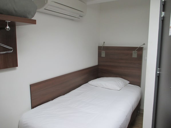 Una habitación pequeña con una cama individual, cabecera de madera, colchón blanco y aire acondicionado de pared.
