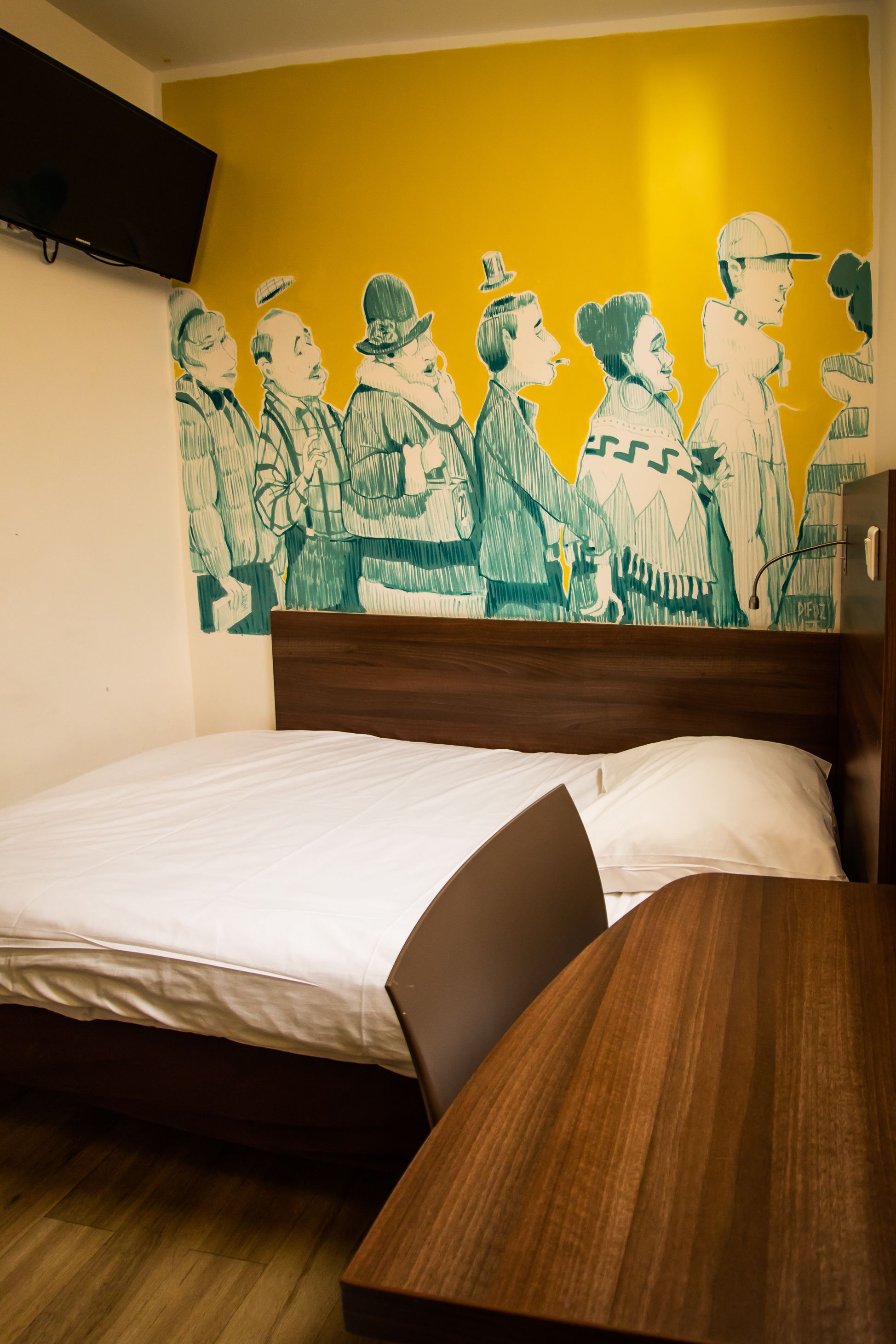 A room with a bed, a wooden bedside table, a wall-mounted TV, and a mural of people.