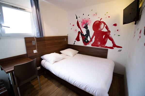 Ein Hotelzimmer mit einem Bett, einem Schreibtisch und einem Stuhl. Es gibt ein Fenster mit Vorhängen, einen an der Wand befestigten Fernseher und ein Wandbild.