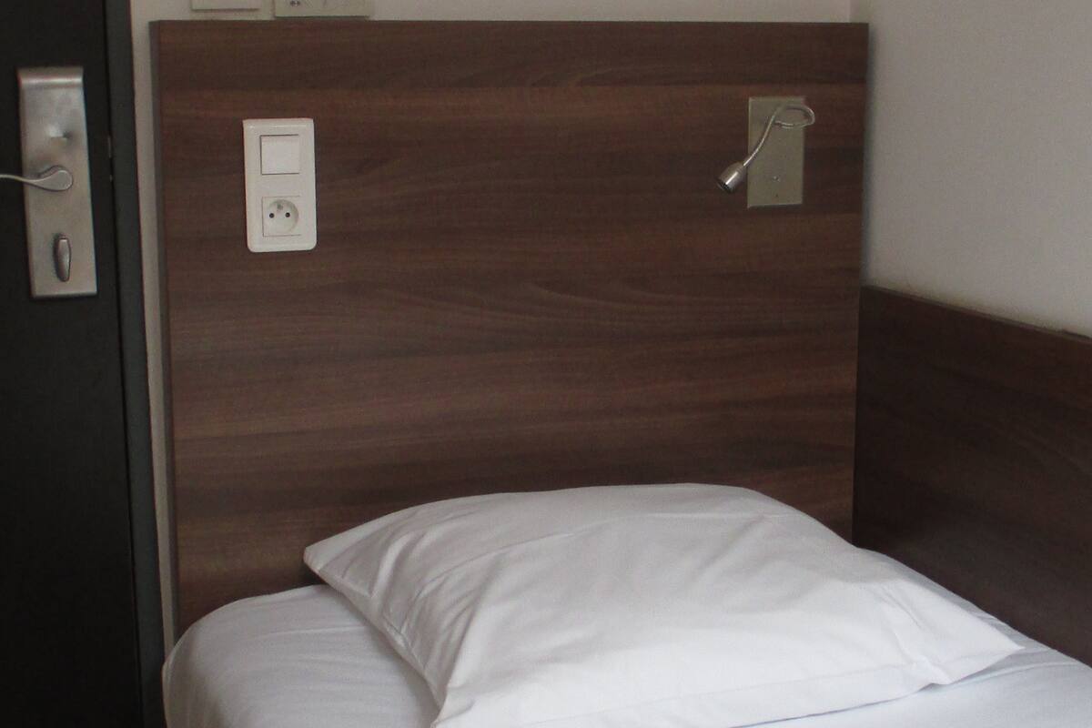 A hotel room with a wooden headboard, a single bed with white bedding, and a wall-mounted light fixture.