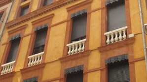 Un edificio de varios pisos con fachada amarilla, ventanas grandes con contraventanas y balcones con barandillas blancas.