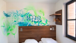 Una habitación con dos camas, un cabecero de madera, una pared con grafitis, un taburete pequeño de madera y una ventana con vistas al exterior.