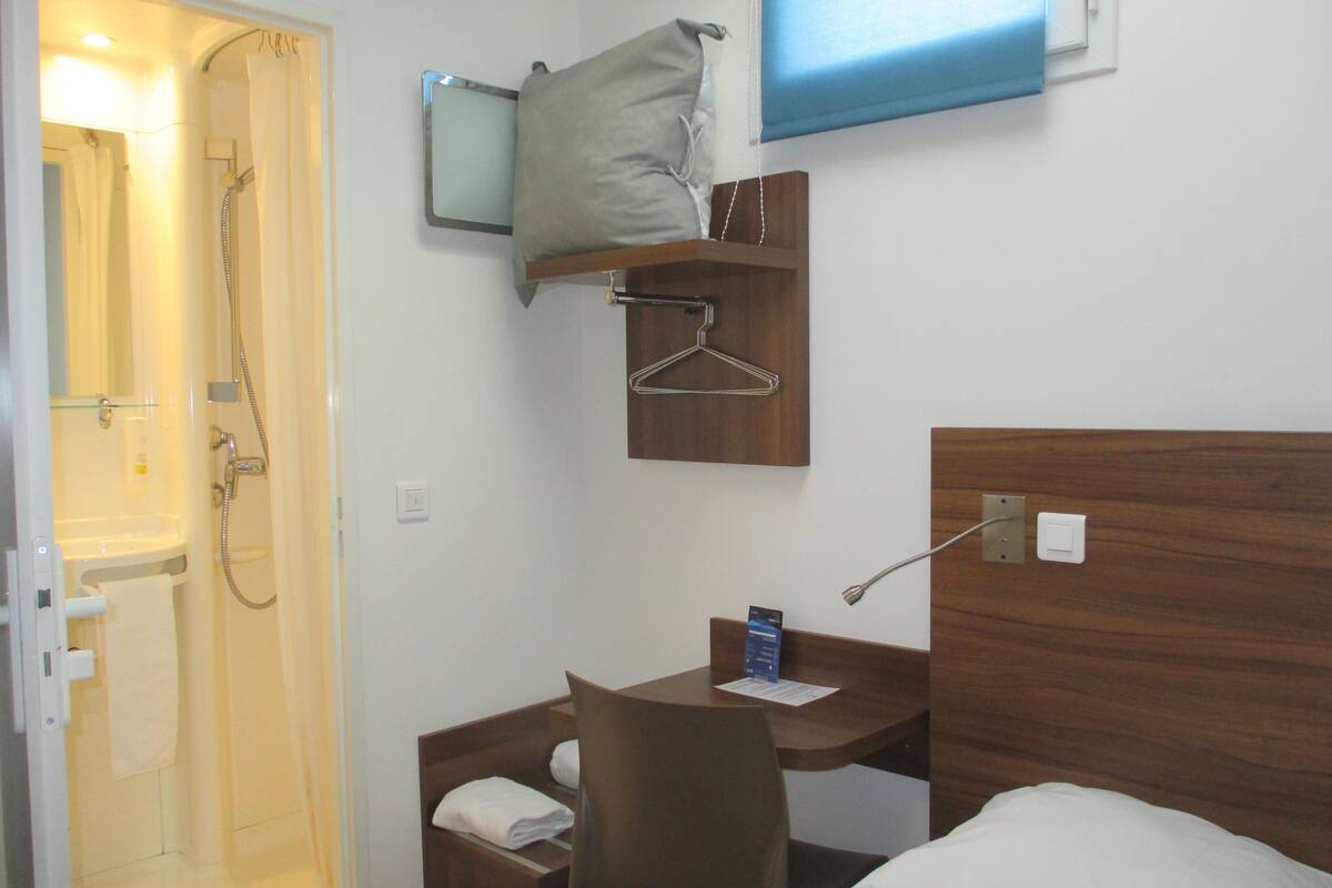 A hotel room with a shower, a desk with a chair, a bed, a TV, and a wooden headboard.