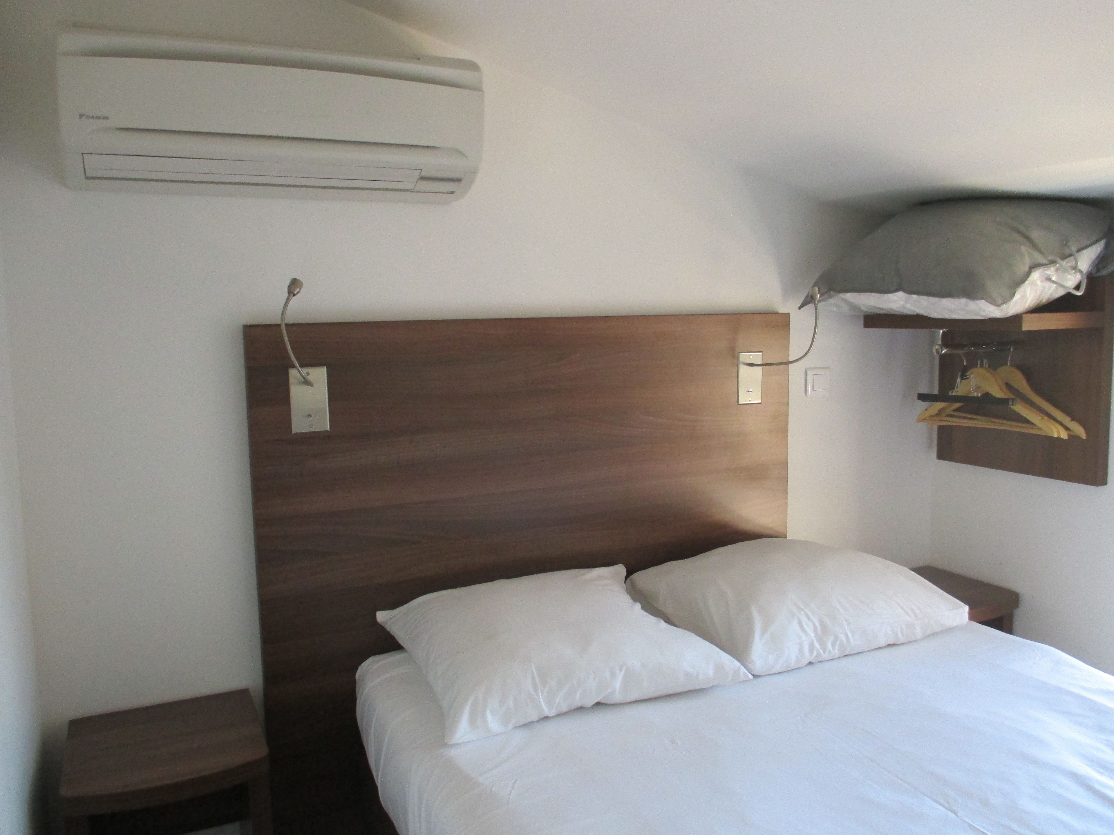 A hotel room with a wooden headboard, a bed with white bedding, a nightstand, a wall-mounted air conditioner, and a coat rack with hangers.