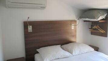 A hotel room with a wooden headboard, a bed with white bedding, a nightstand, a wall-mounted air conditioner, and a coat rack with hangers.