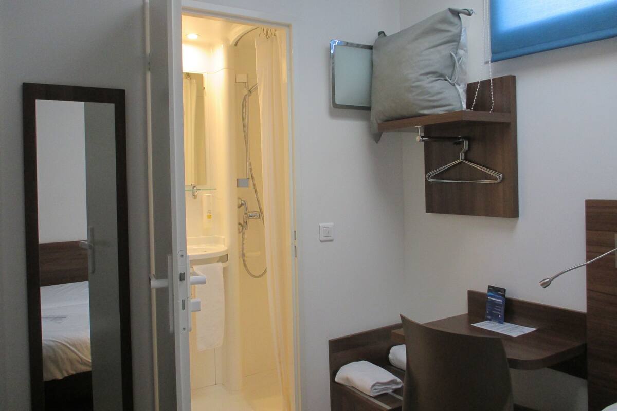 A hotel bathroom with a shower, a sink, a mirror, a towel rack, and a small table with a chair.