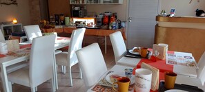 A dining area with white chairs and tables, a kitchen in the background, and a reception desk. - La Suite di Segesta (Calatafimi)