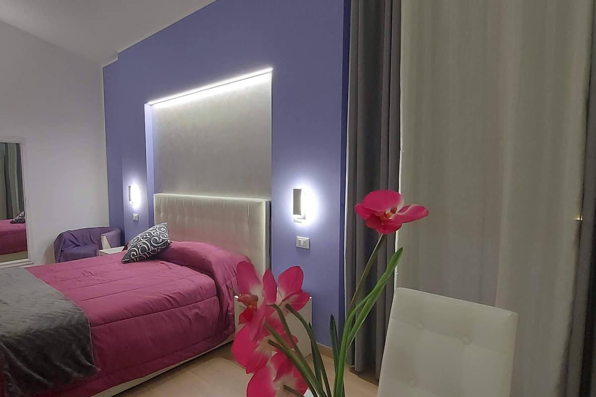 Quarto moderno com cama grande, mesa de cabeceira, cadeira, espelho e janela com cortinas.